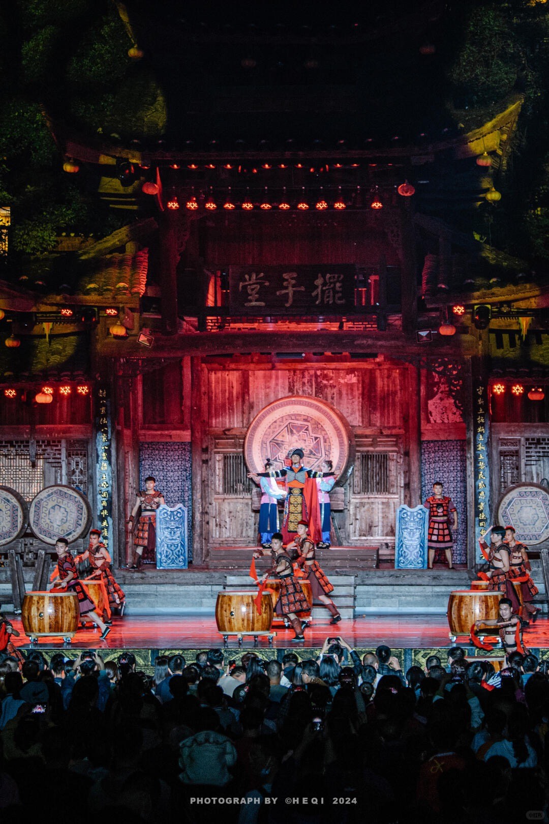 Costumed performers with barrel drums on a lantern-lit wooden stage before a large night-time audience.