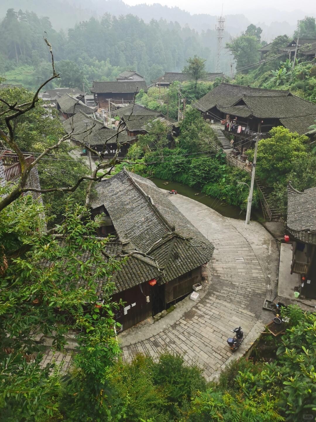 Cluster of grey-tiled timber village houses beside a paved lane and small stream below misty forested hills.