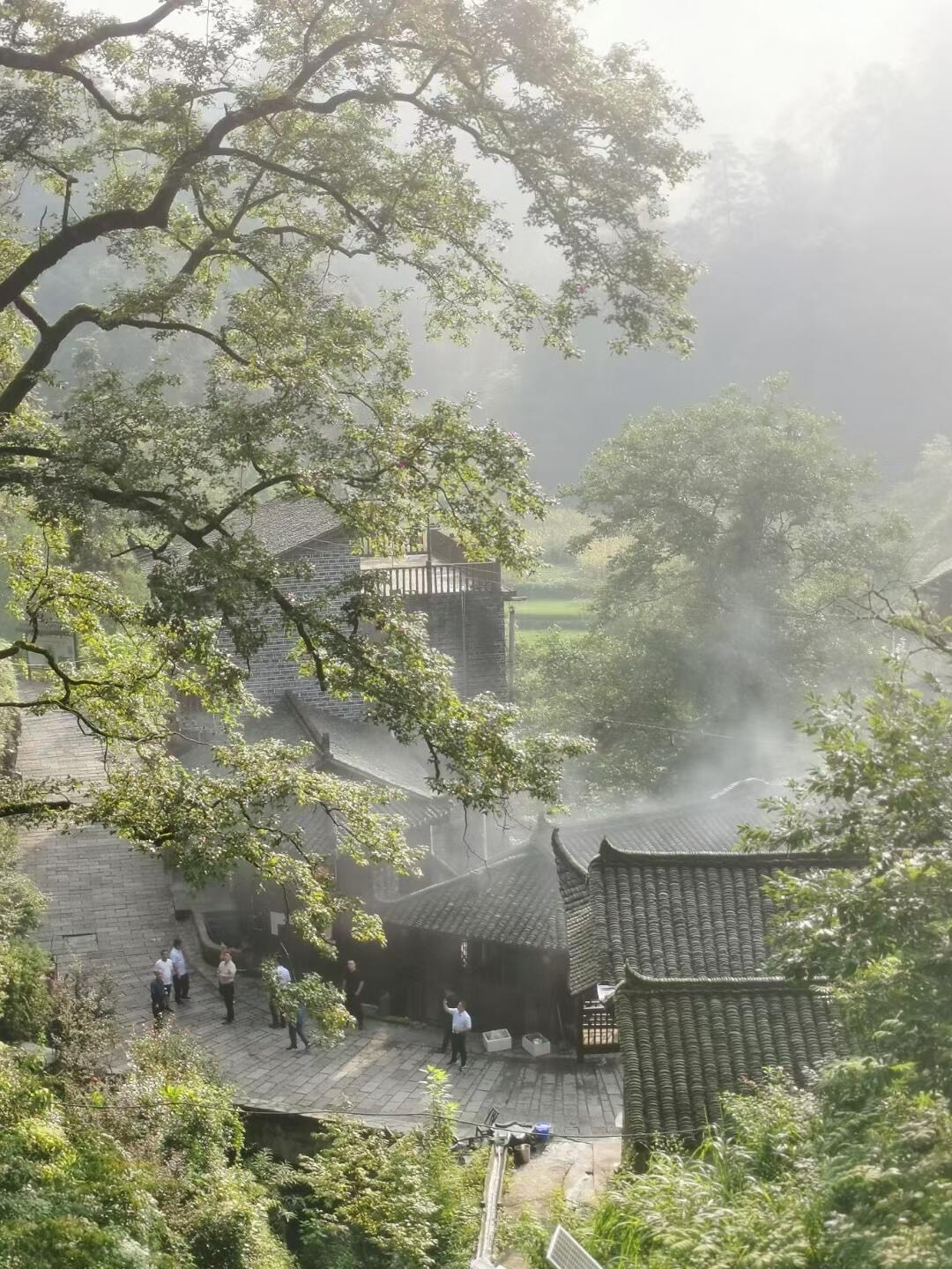 Grey-tiled traditional courtyard houses among trees with people gathered on a stone terrace in soft morning mist.
