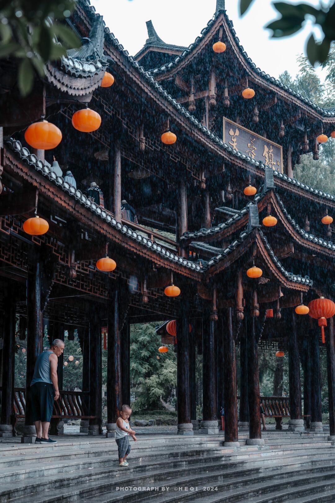 Tiered wooden pavilion hung with orange lanterns in the rain as an elderly man watches a toddler on the steps.