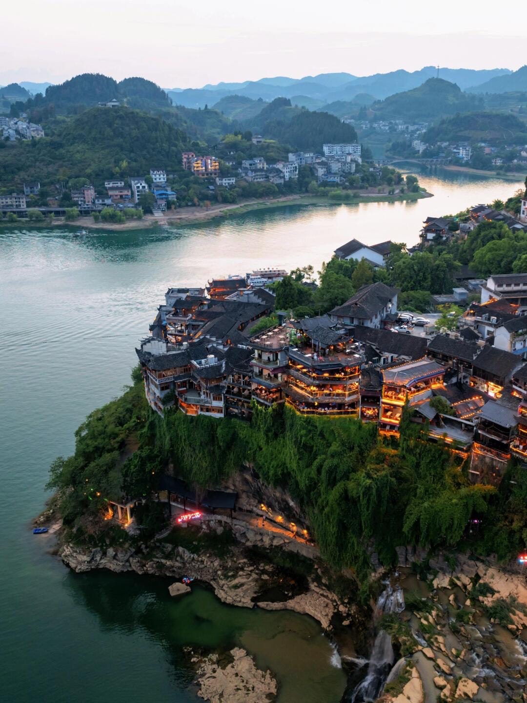 Aerial dusk view of a riverside old town, its lantern-lit timber houses clustered on a green clifftop above a bend in the river.
