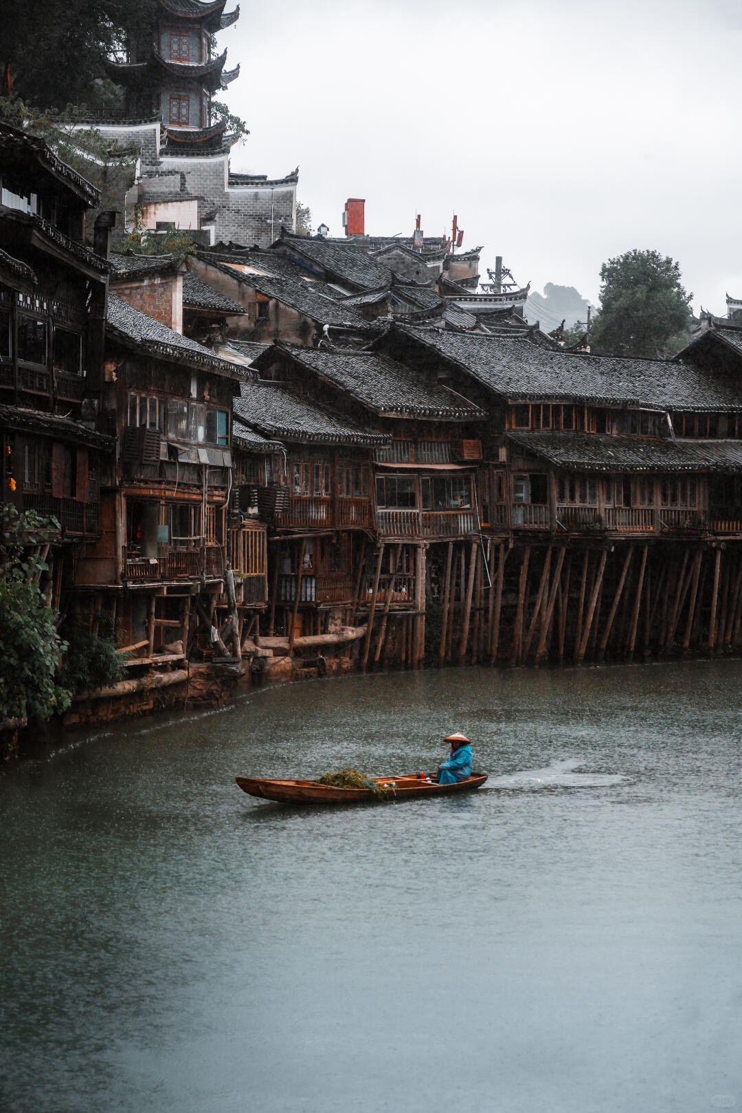 A boatman in a straw hat and blue raincoat rows a wooden boat past riverside wooden stilt houses in the rain.