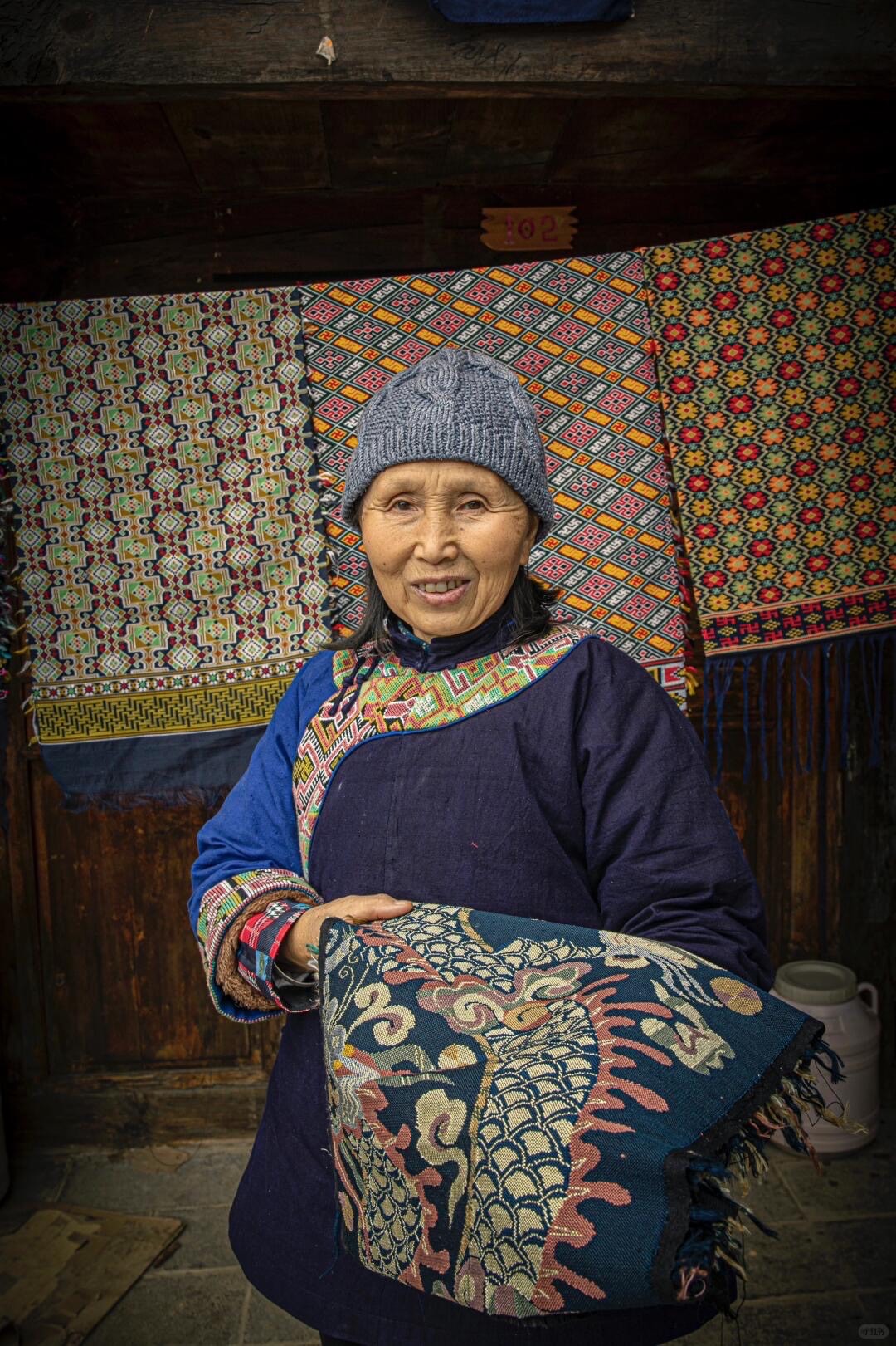 Smiling elder woman in indigo embroidered dress holds a dragon-patterned brocade in front of hanging woven textiles.
