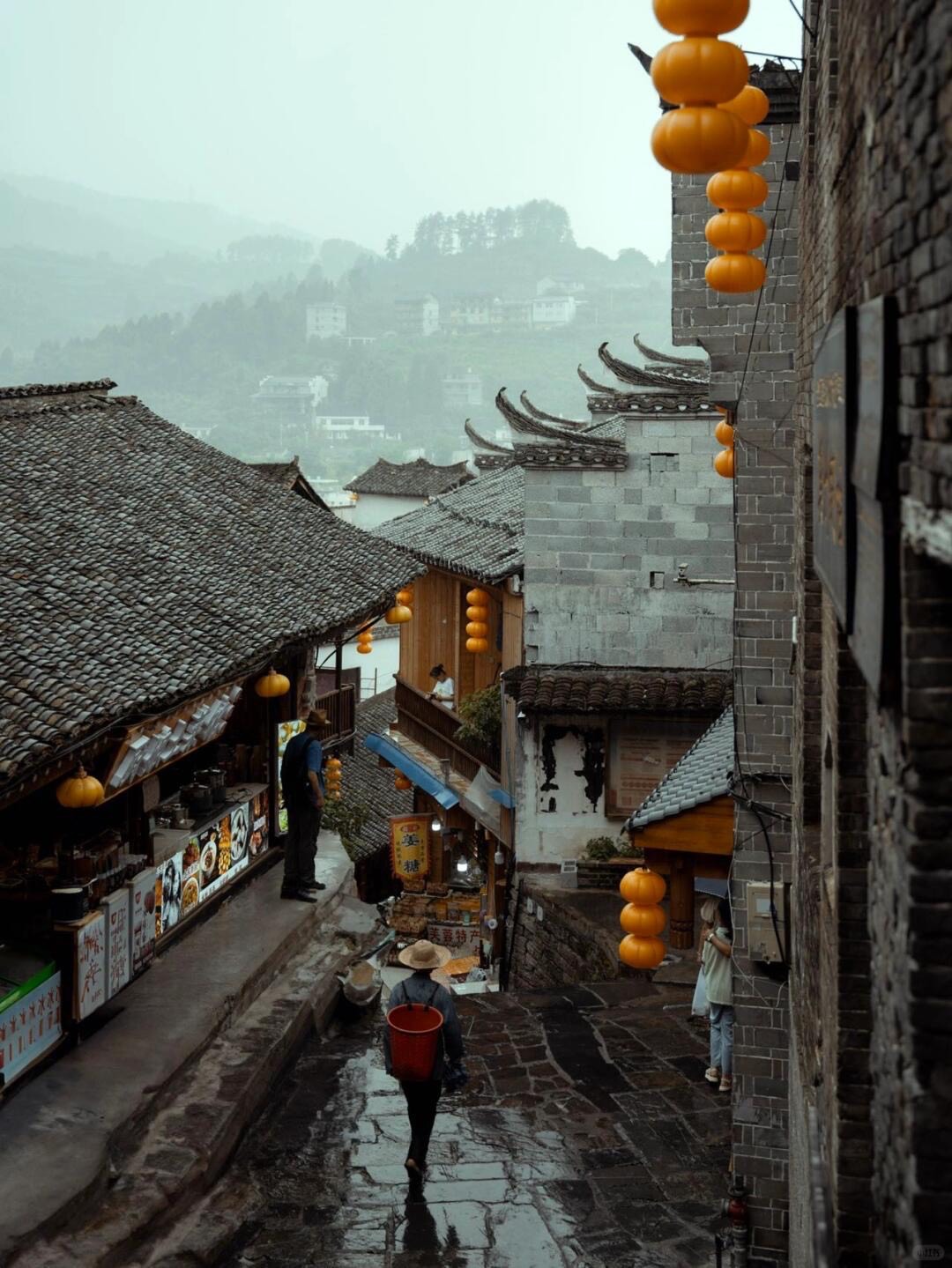 Rain-soaked stone alley lined with tiled-roof shops and orange lanterns as a porter walks downhill past hillside houses.
