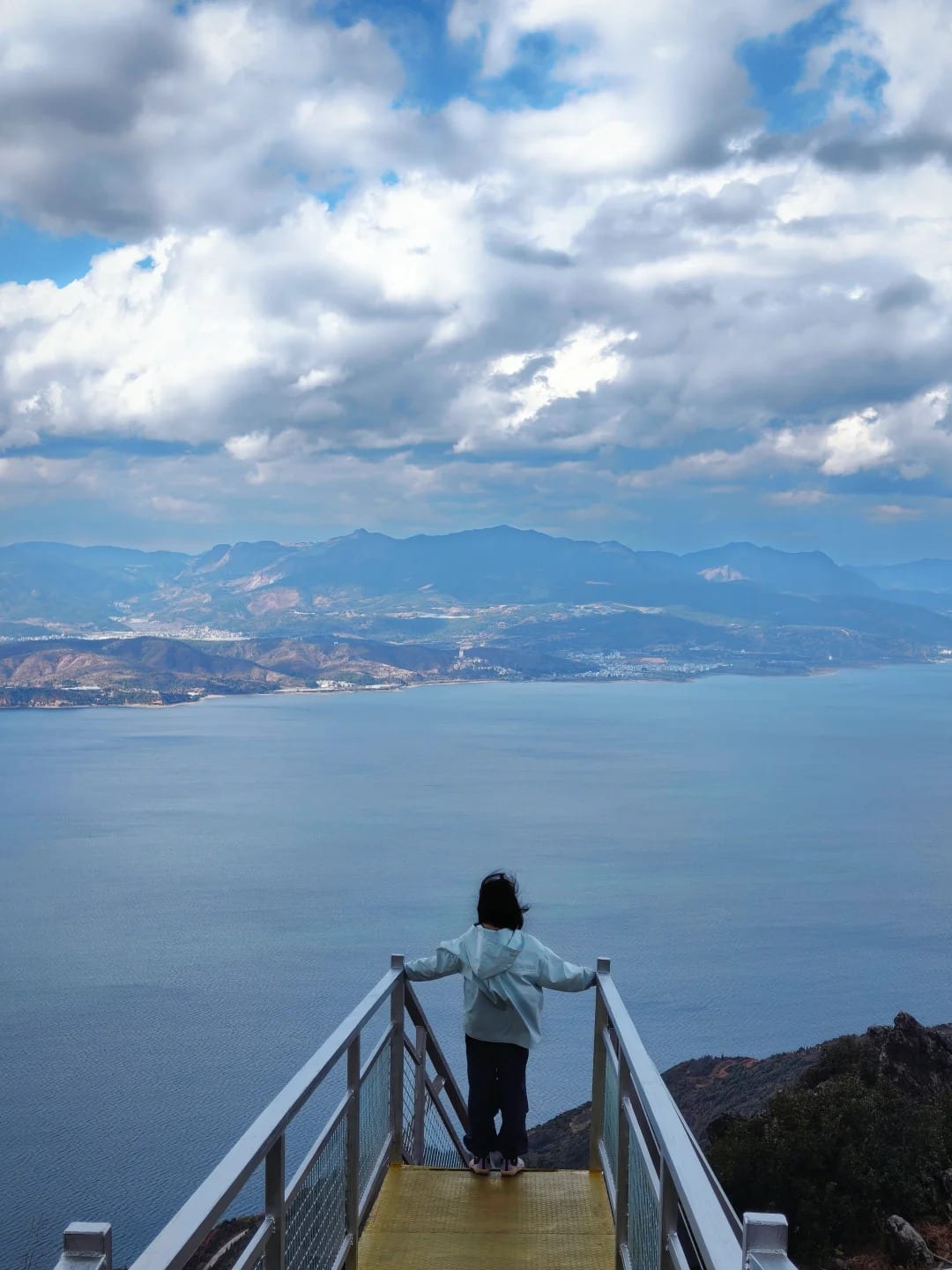 A walker at the railing of a viewing platform on the cliff boardwalk, Fuxian Lake spread out below under a big sky