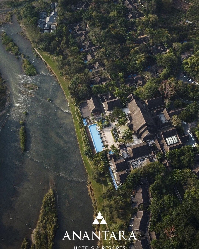Aerial view of Anantara Xishuangbanna's swimming pool and thatched villas set among trees beside a winding river.