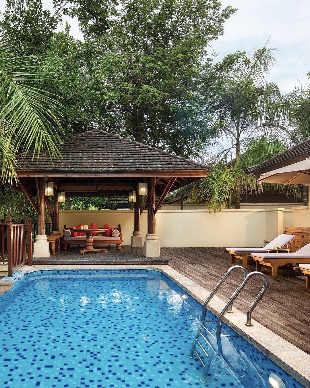 Private mosaic-tiled plunge pool beside a timber sala with daybed and sun loungers framed by tropical palms.