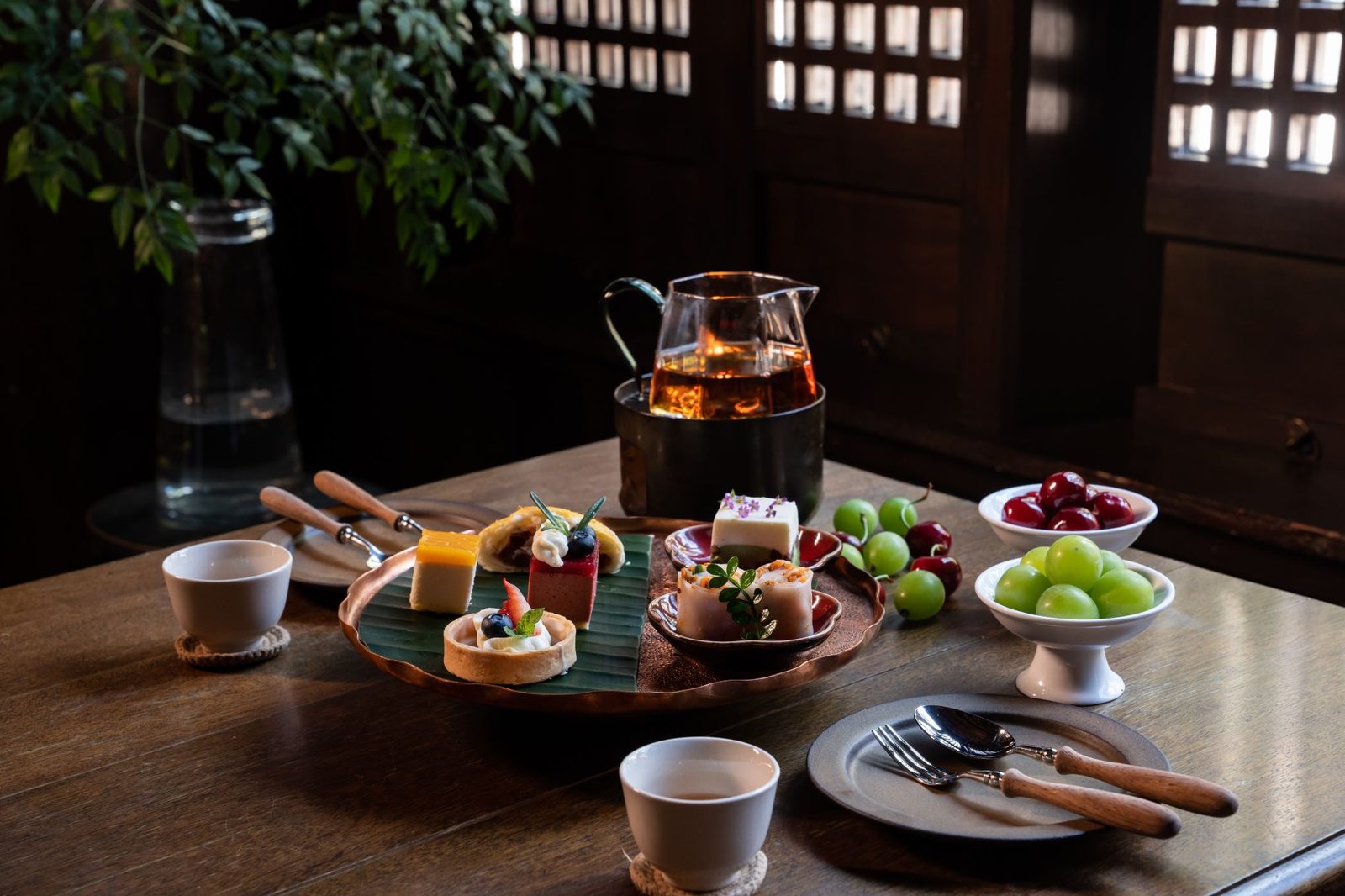 Afternoon tea with a glass teapot, small cakes and fruit on a timber table.