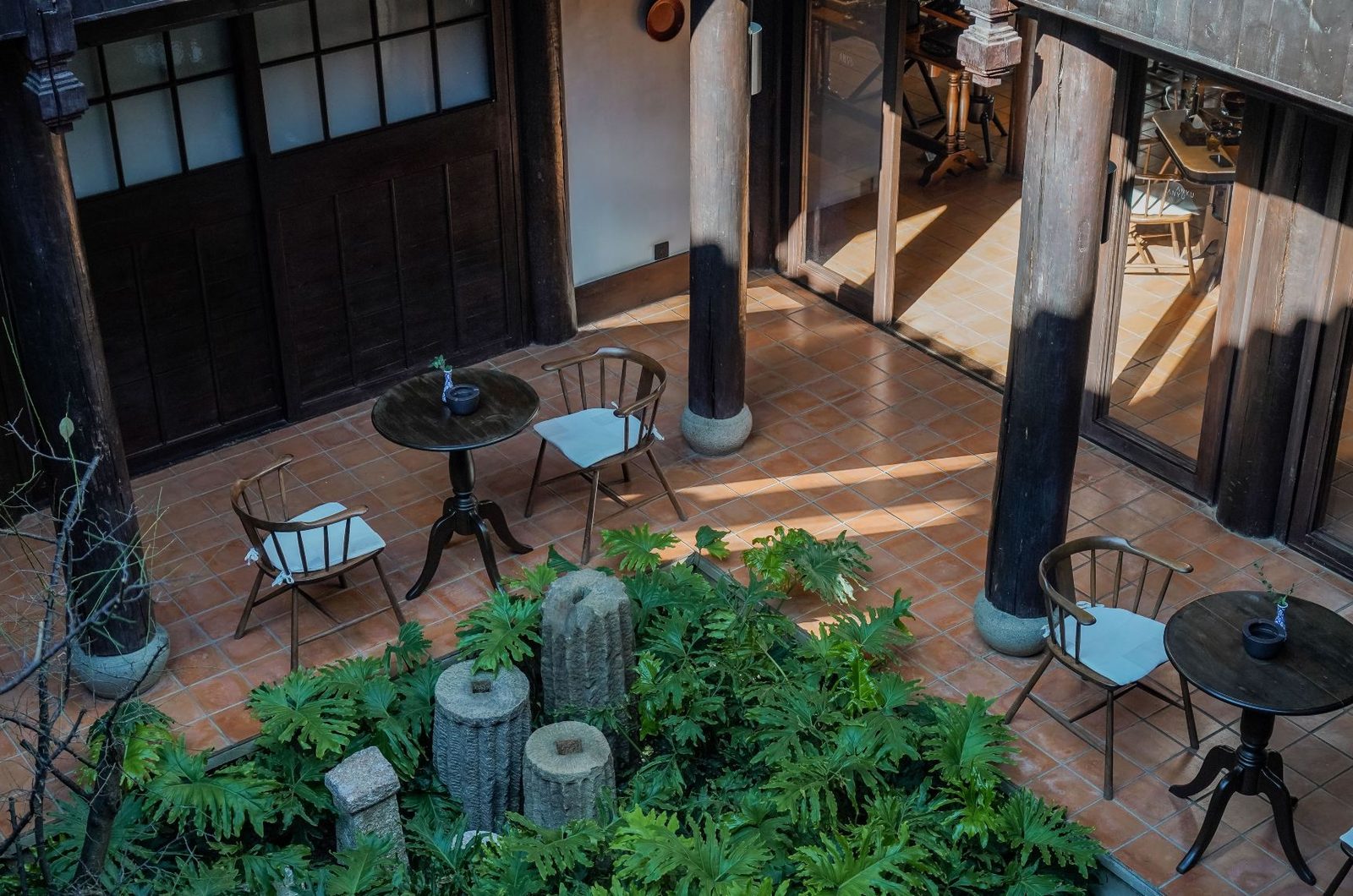 Sunlit courtyard terrace with café chairs and small round tables on terracotta tiles, ferns in the foreground.