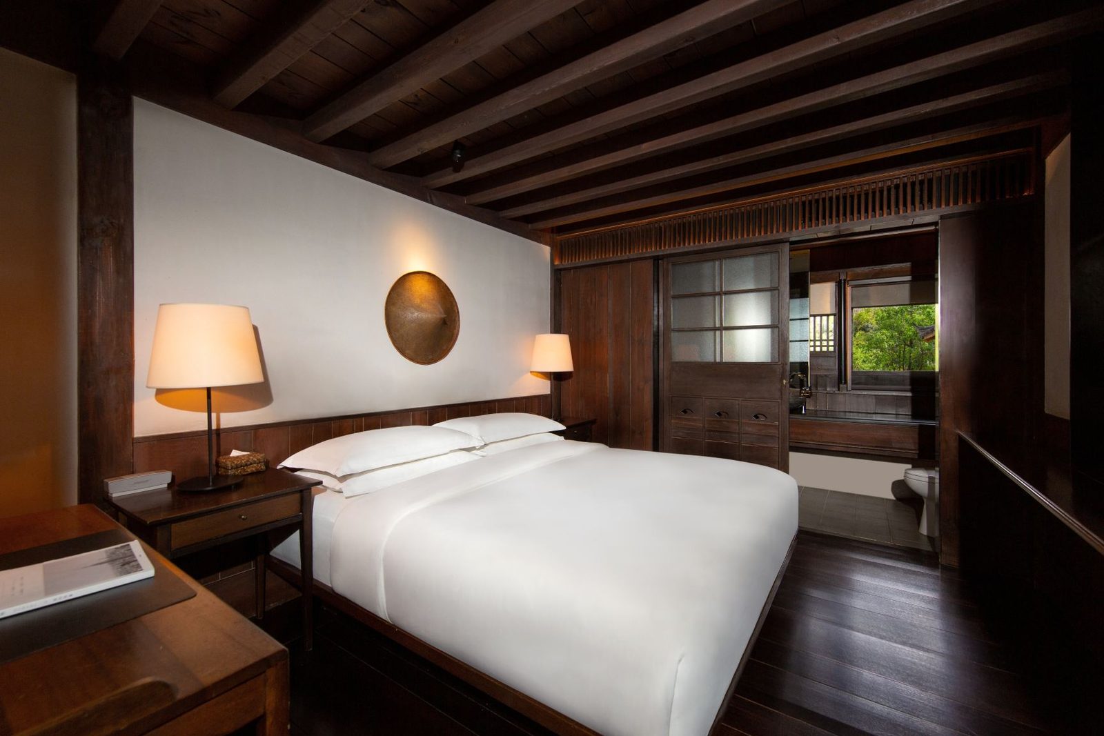 King room in warm timber with a round bronze mirror, bedside lamps and a window onto greenery.
