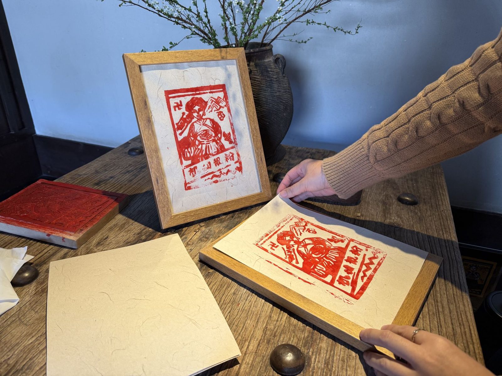A guest lifting a freshly pulled red woodblock print, a framed print standing on the table behind.