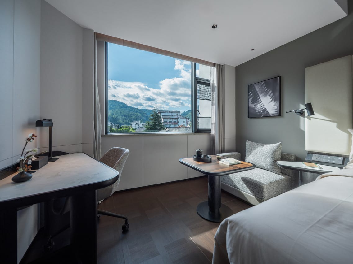Atour Hotel, Zhangjiajie Forest Park Gate — a writing desk, tea table and reading chair beside a window onto green hills and white-walled houses