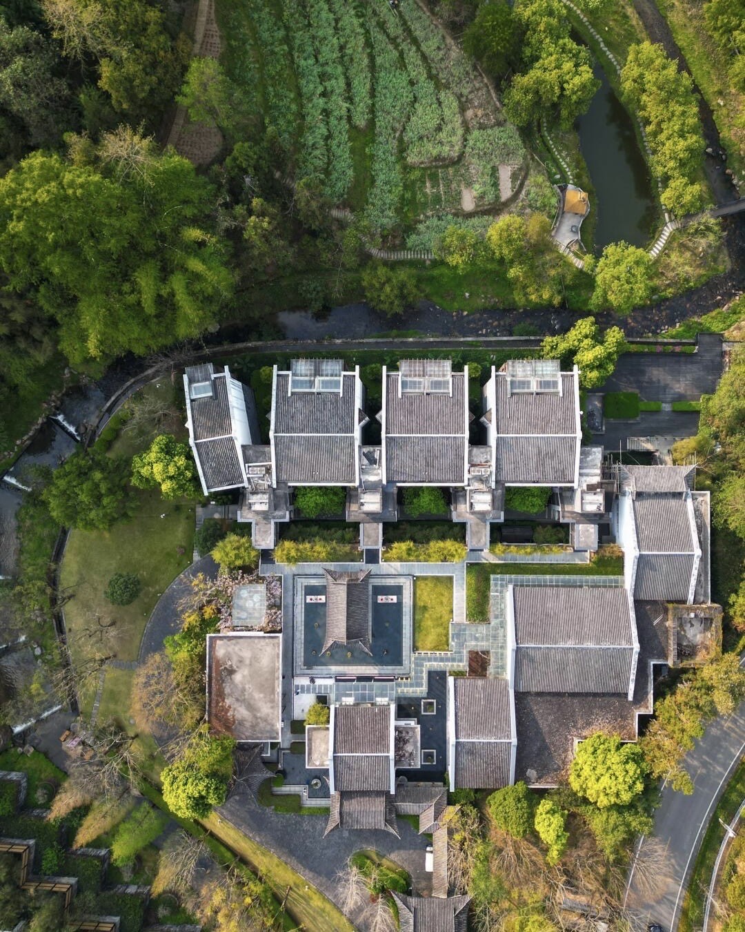 Aerial view of grey tiled-roof courtyard villas set among terraced fields, streams and dense green forest.