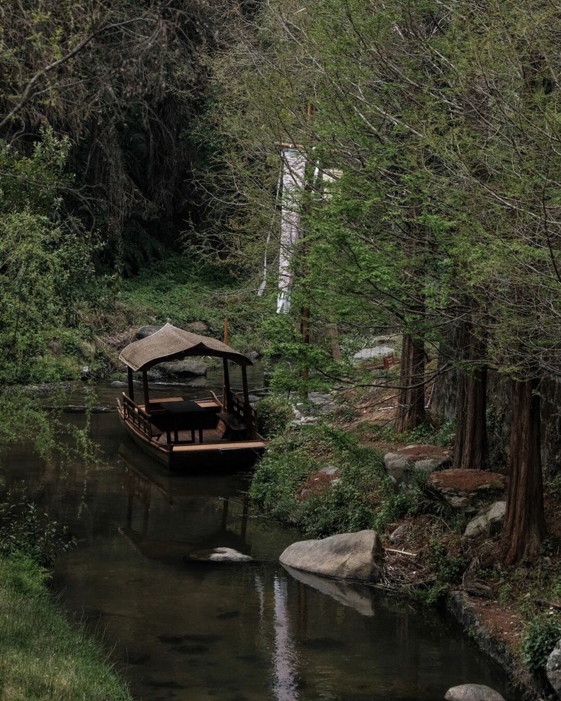 Canopied wooden boat moored on a calm forested stream lined with rocks and slender trees.