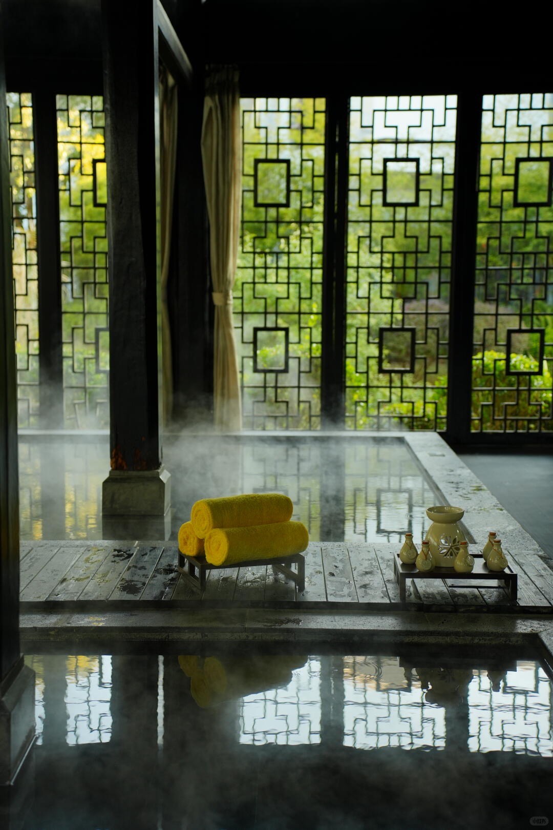 Steaming indoor spa pool beside a timber deck with rolled yellow towels and ceramic bottles, framed by geometric lattice screens.