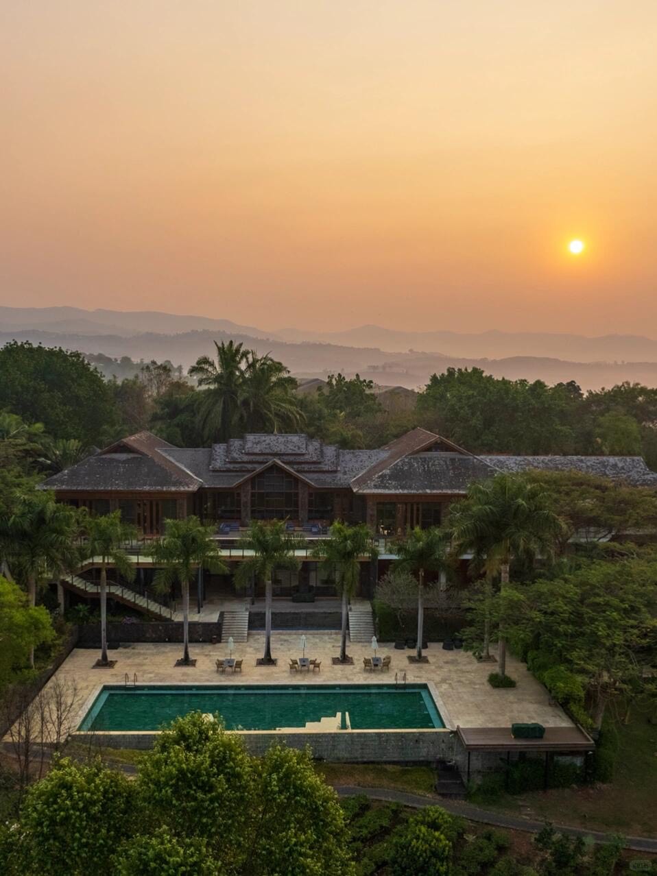 Aerial view of a timber resort and rectangular pool at sunset, with hazy layered hills fading into the distance.