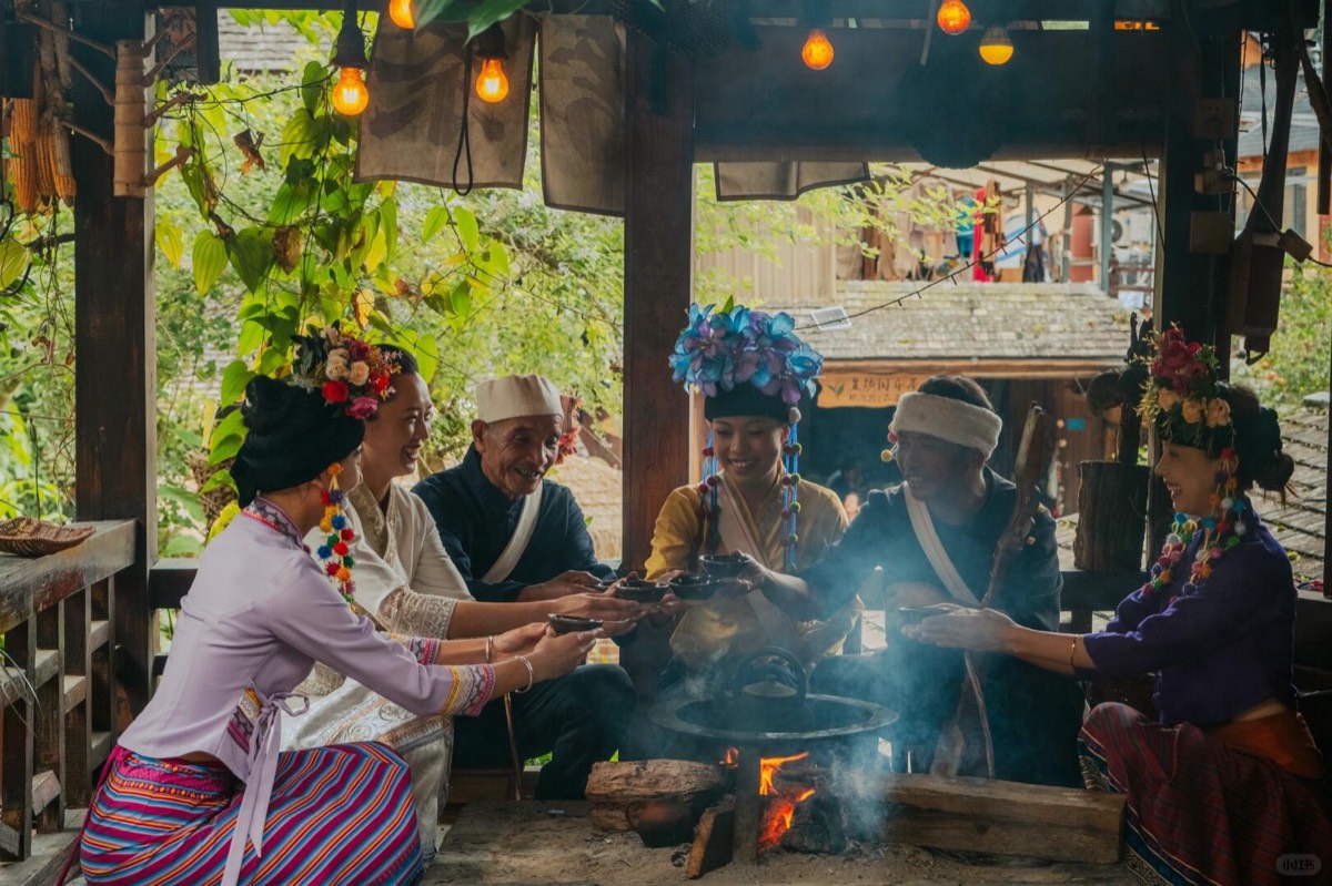 People in floral ethnic headdresses raise tea cups around a smoking firepit inside a rustic timber pavilion.