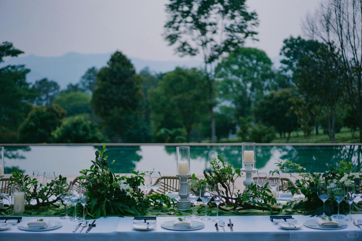 Long candlelit dinner table dressed with greenery beside a reflecting pool, mountains hazy beyond.