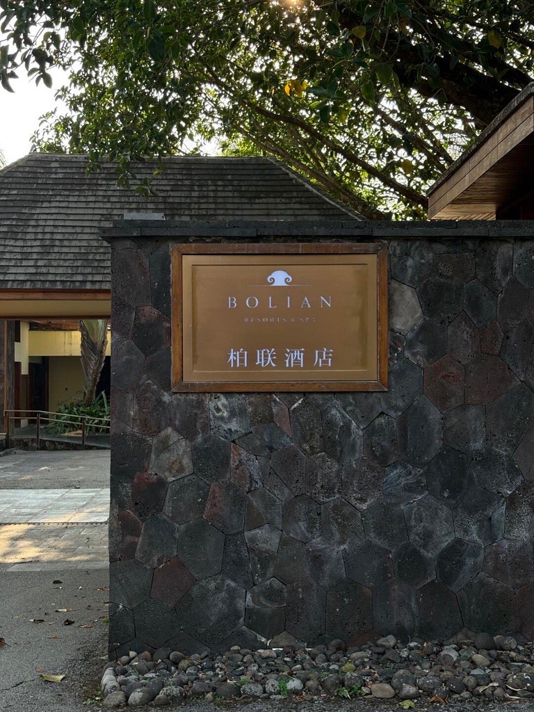 Bolian resort entrance sign mounted on a dark volcanic-stone wall beneath overhanging trees.