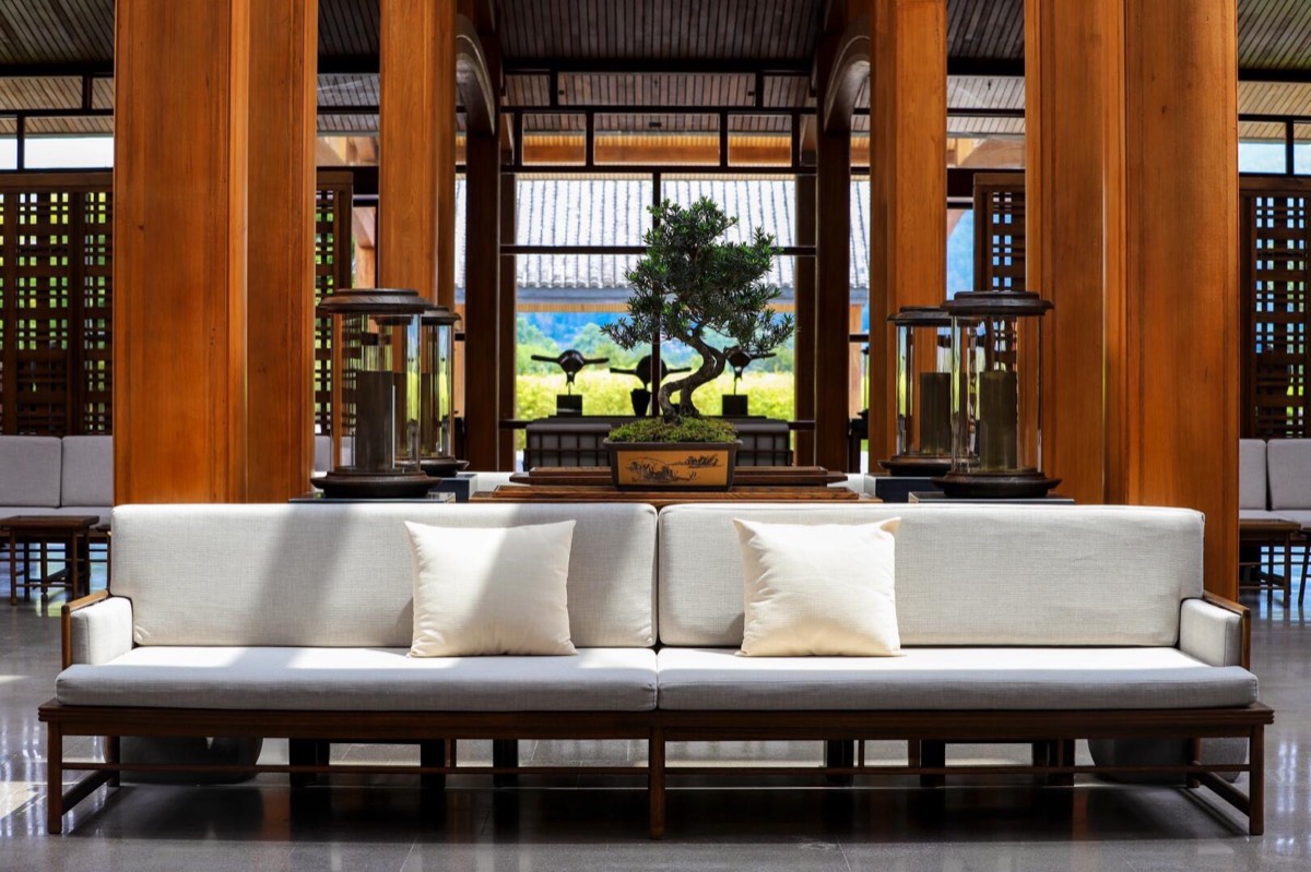 White cushioned sofa flanked by metal lanterns and a bonsai tree among tall wooden columns.