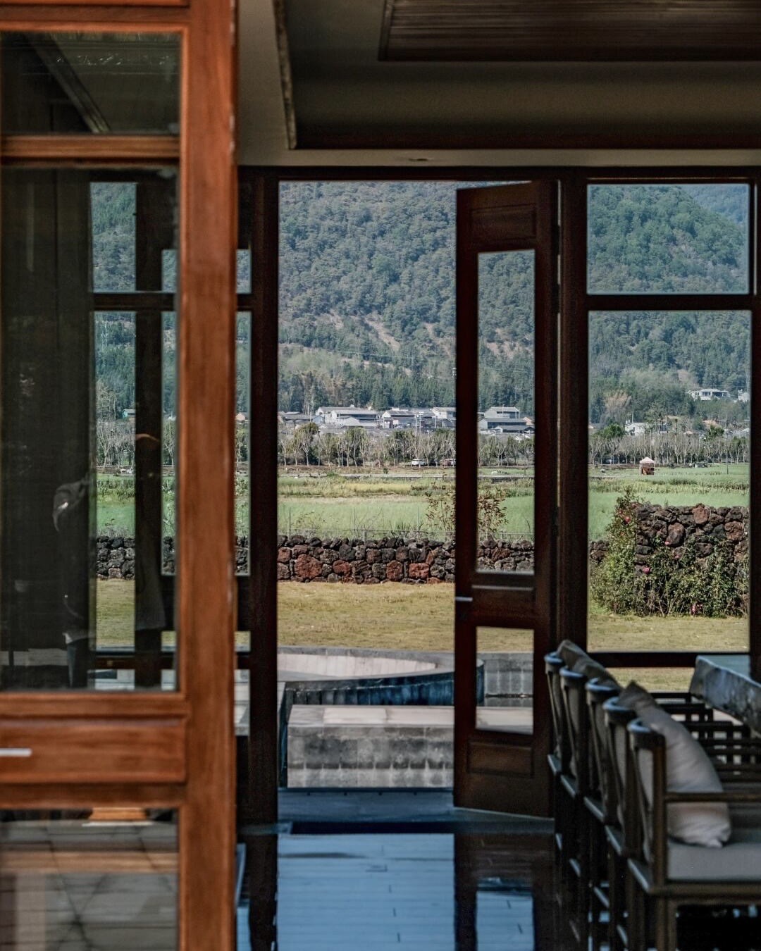 Open timber doors framing a view of farmland, a stone wall and forested mountains beyond a terrace.