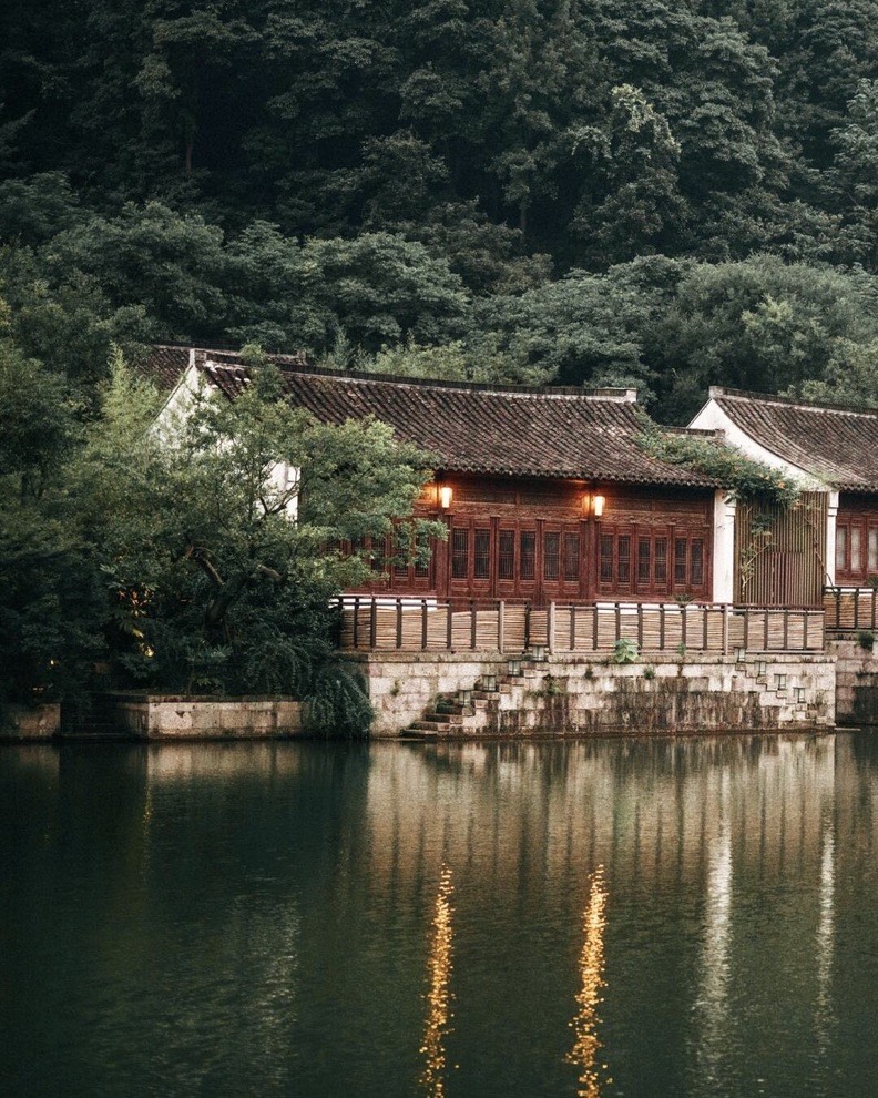 Lantern-lit wooden Chinese pavilion reflected in a still lake below a forested hillside at dusk.
