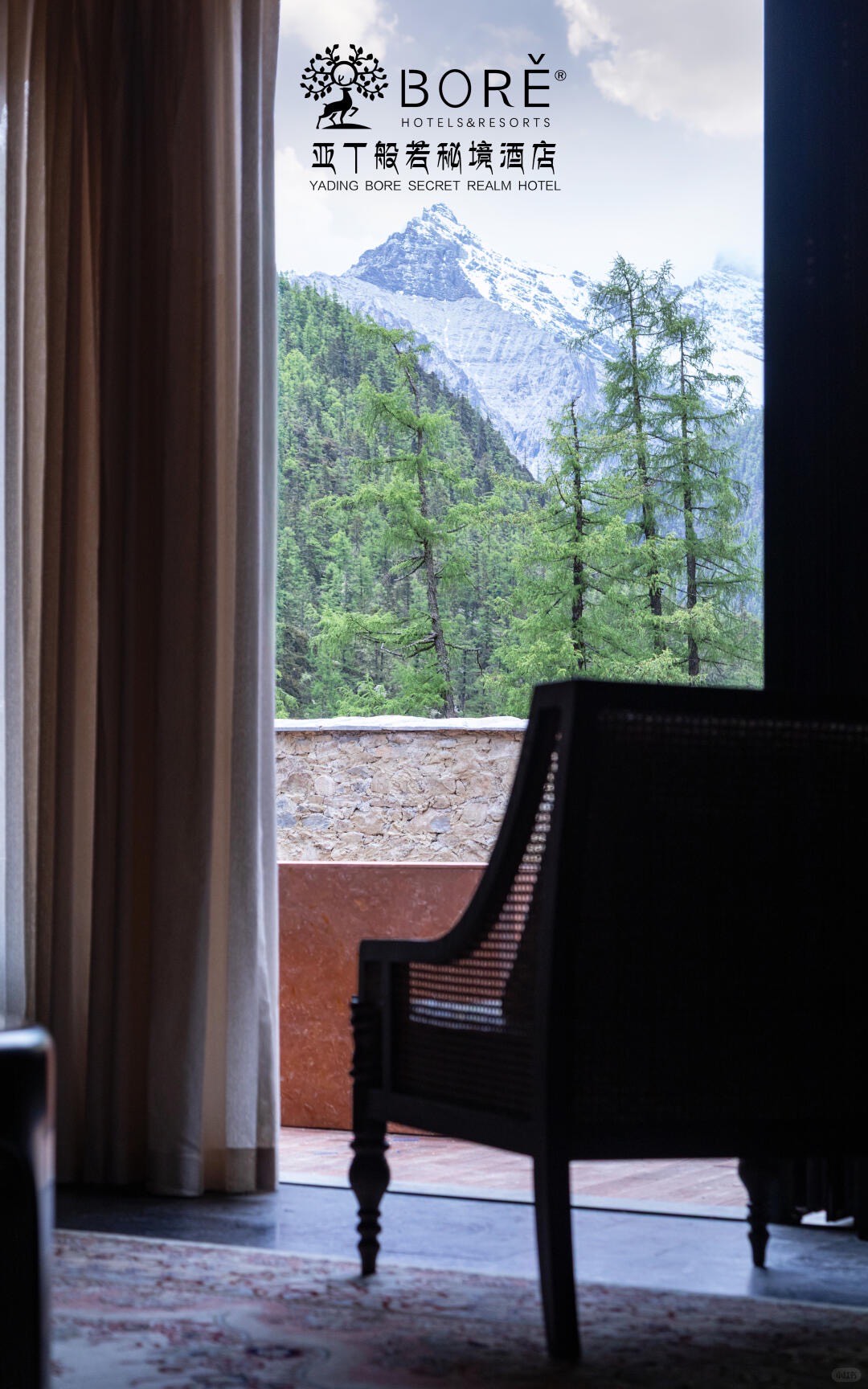 Guestroom armchair beside a curtained window framing pine forest and a snow-capped peak at Bore Yading.