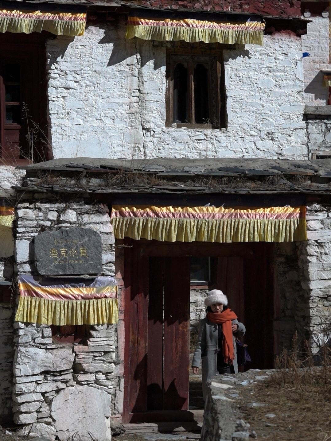 Visitor stepping through the red doorway of a whitewashed Tibetan stone building draped with yellow prayer banners at Bore Yading.