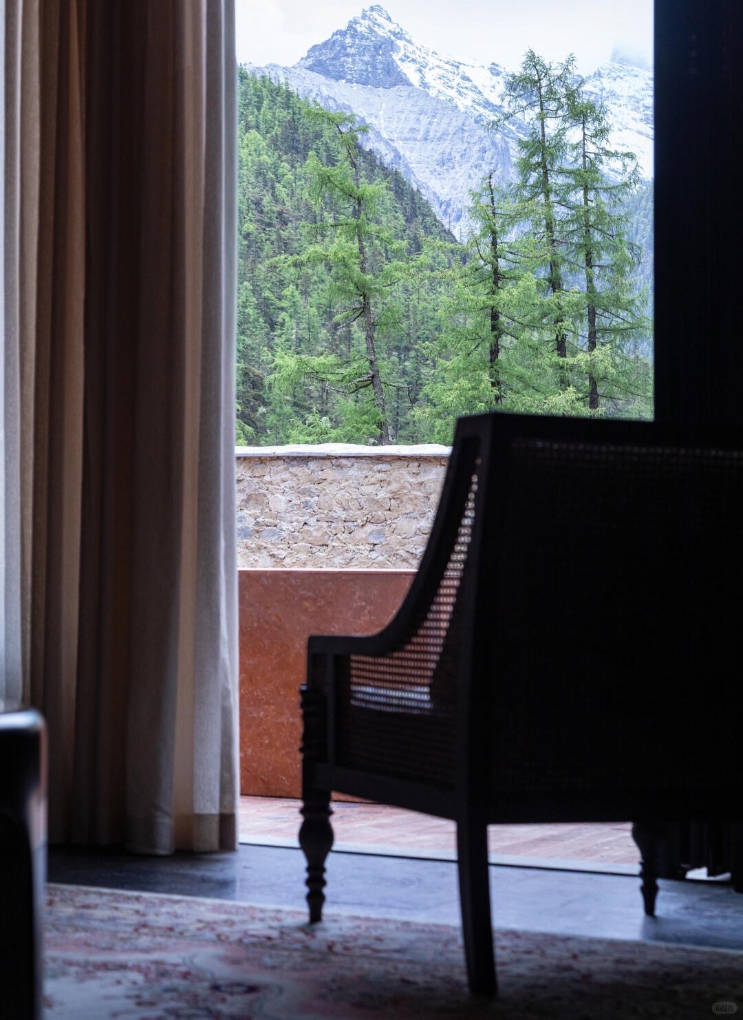 Guestroom armchair beside a curtained window framing pine forest and a snow-capped peak at Bore Yading.