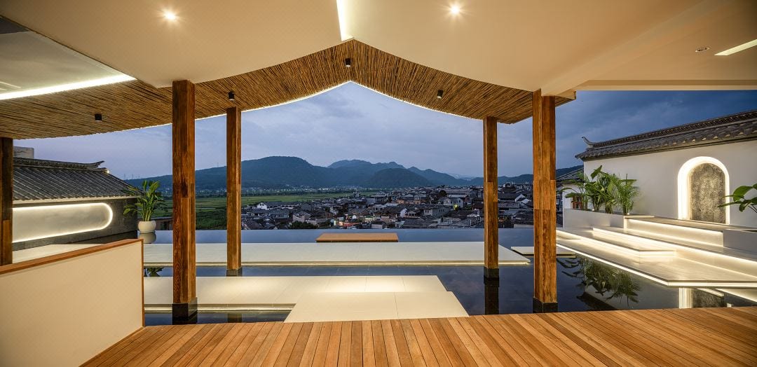 Boyun Hot Spring Inn — an open timber-and-thatch pavilion with a still reflecting pool, framing Heshun's tiled rooftops and the far mountains at blue hour