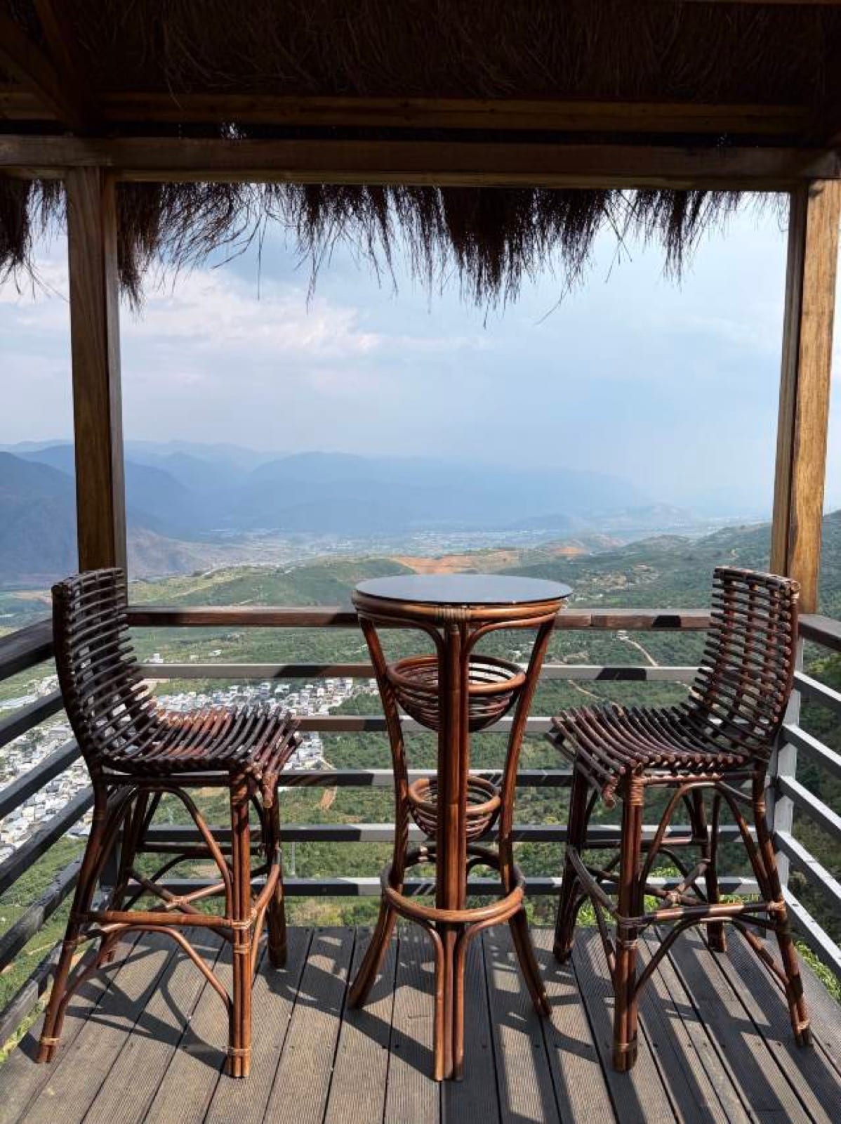 Huke Coffee Manor — a thatched cliff-edge deck with a rattan table and stools, looking down over villages and the hazy mountains of the Nujiang valley