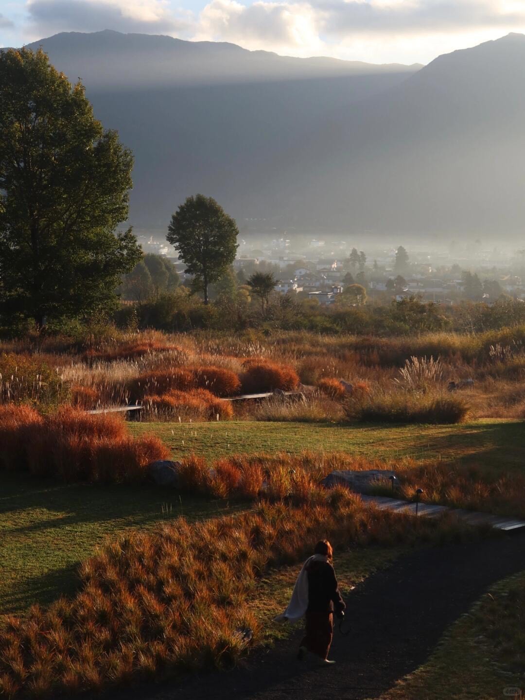 Guest walking a garden path through russet ornamental grasses above a misty valley town at sunrise.