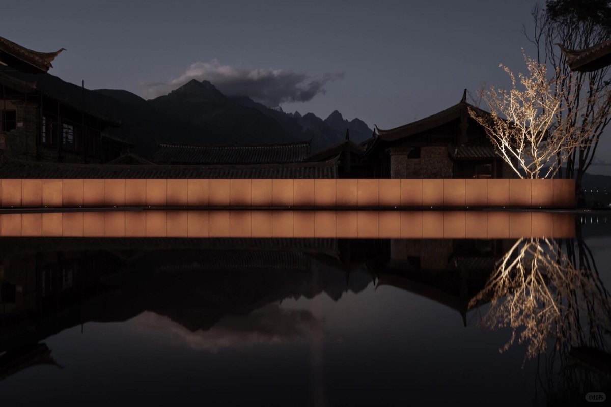 Glowing amber-lit wall mirrored in a still reflecting pool with tiled-roof buildings and jagged mountains at dusk.