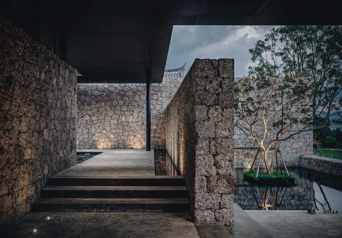 Rough stone-walled courtyard with steps leading past a dark reflecting pool and a slender tree at twilight.