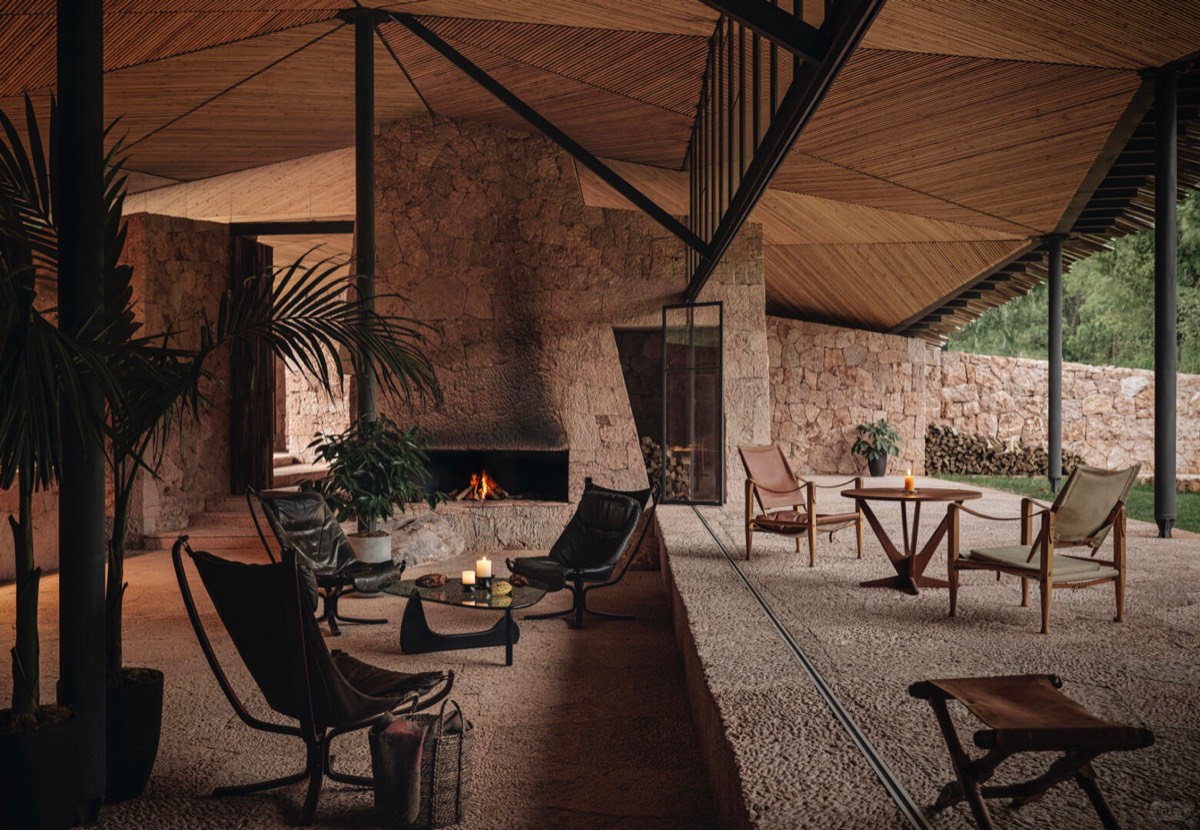 Open-air stone lounge with leather chairs around a lit fireplace beneath a folded timber-clad canopy roof.