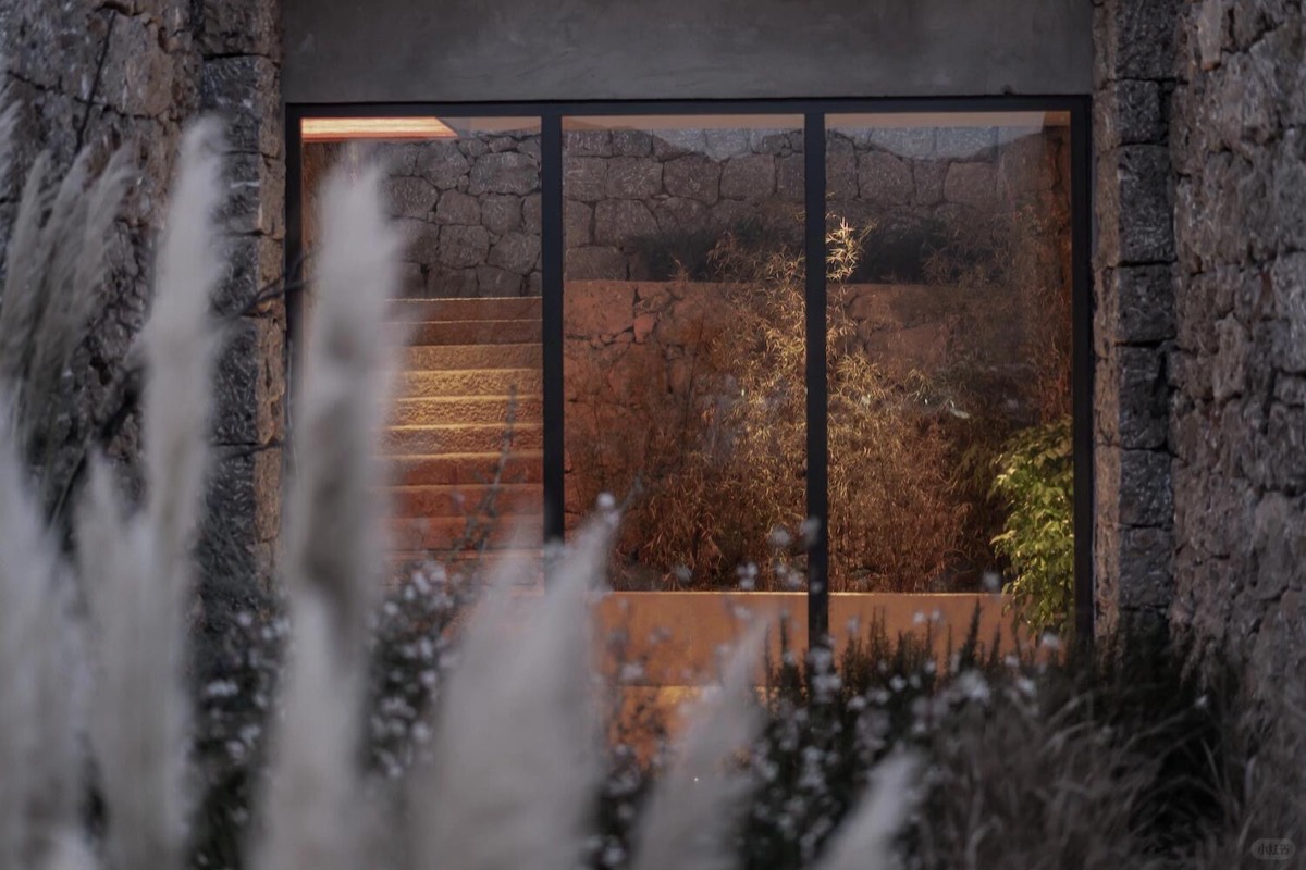 View through a tall window to lamp-lit stone steps and autumn garden foliage, framed by feathery pampas grass.