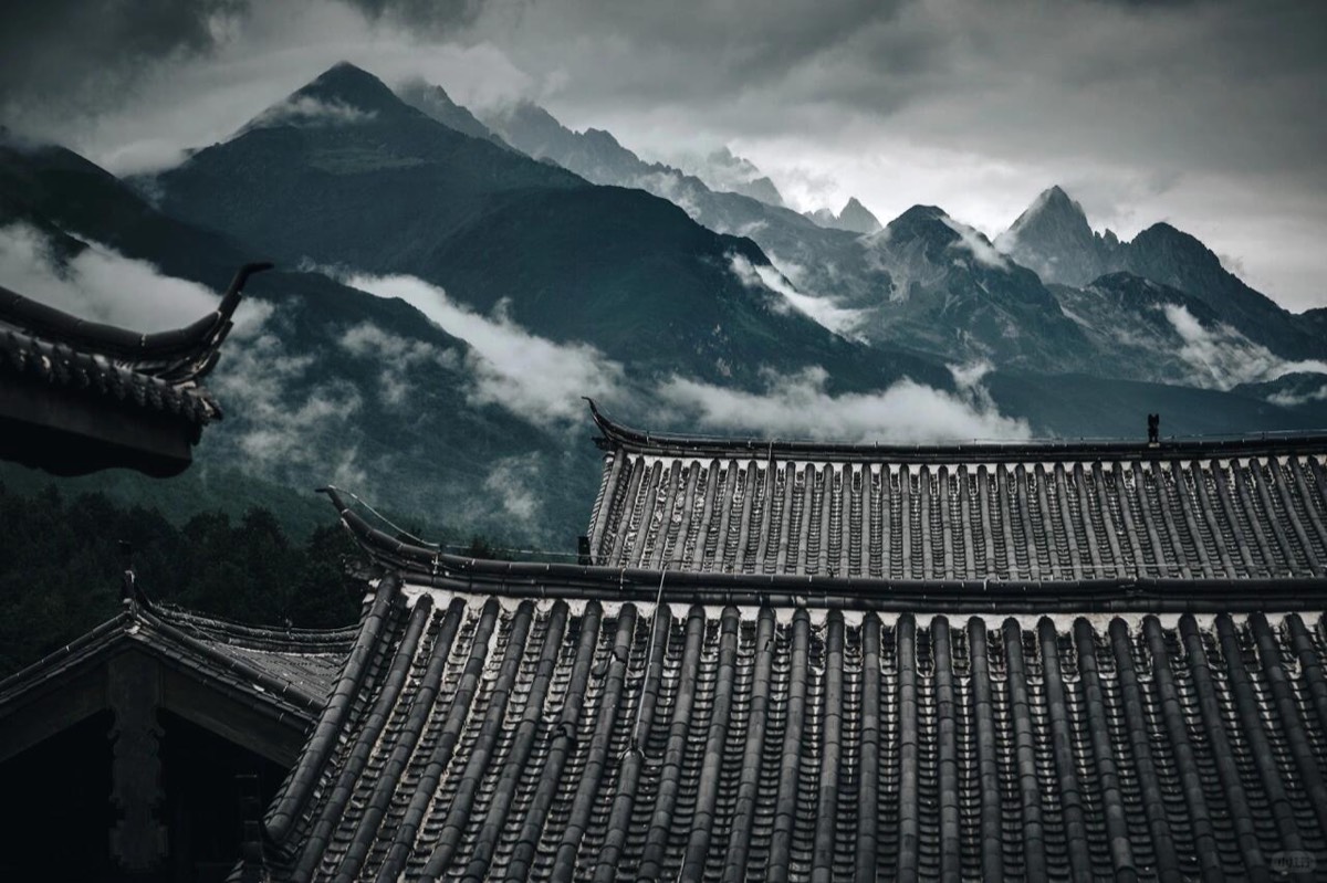 Grey tiled traditional rooftops layered against cloud-wrapped mountain peaks in moody overcast light.