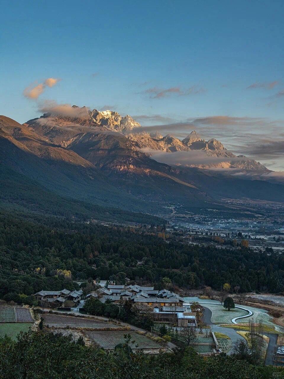 Cluster of tiled-roof hotel buildings in a forested valley below sunlit snow-capped mountains at dawn.