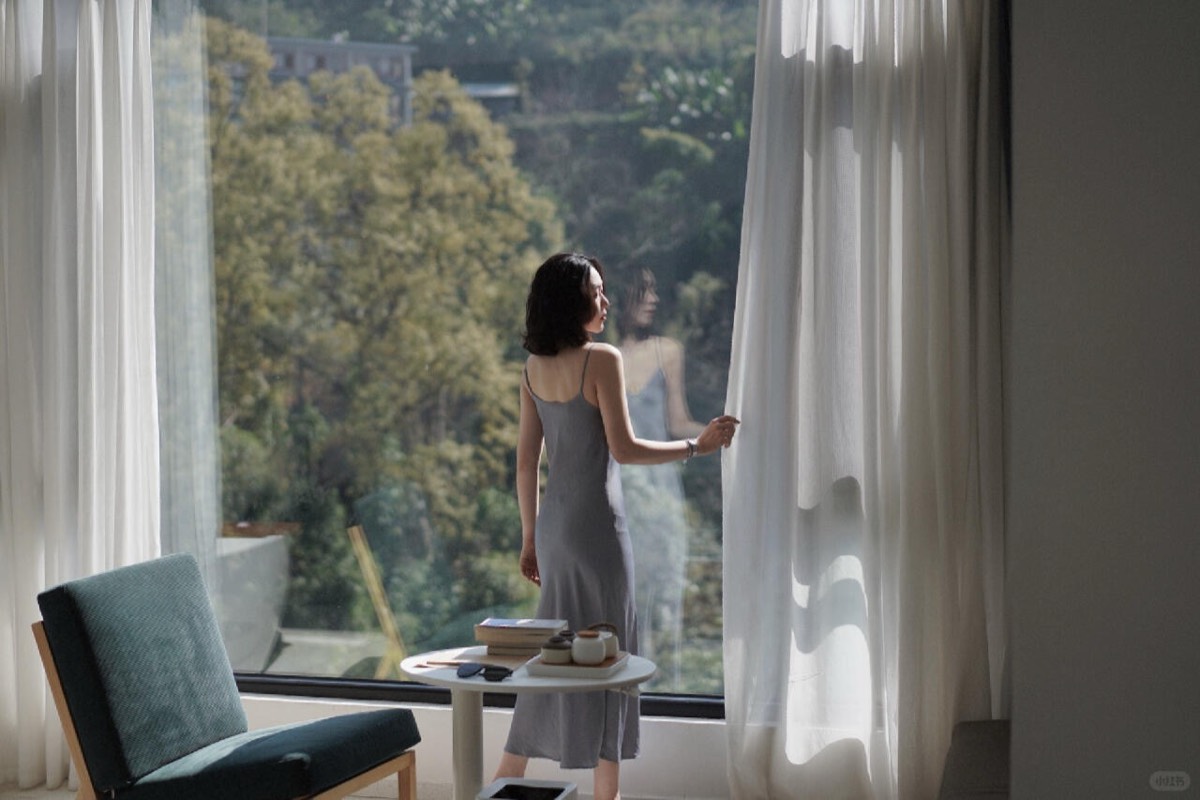 Guest in a grey slip dress drawing a sheer curtain to a floor-to-ceiling window over wooded hills.