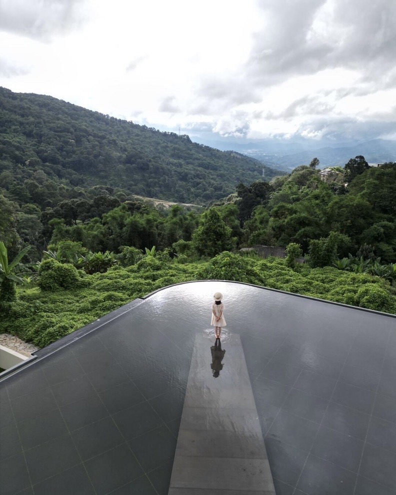 Woman in a white dress standing on a walkway across a dark infinity pool overlooking lush forested hills.