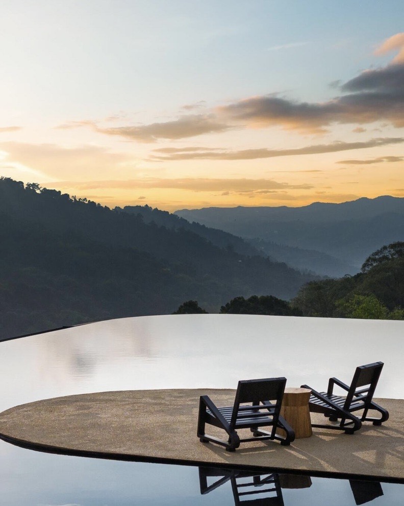 Two low loungers on a stone deck beside an infinity pool facing layered mountain ridges at golden sunset.