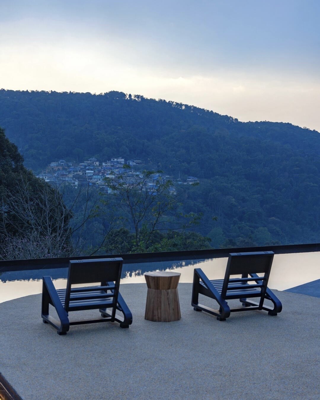 Two black loungers beside an infinity pool overlooking a forested valley and hillside village at dusk.
