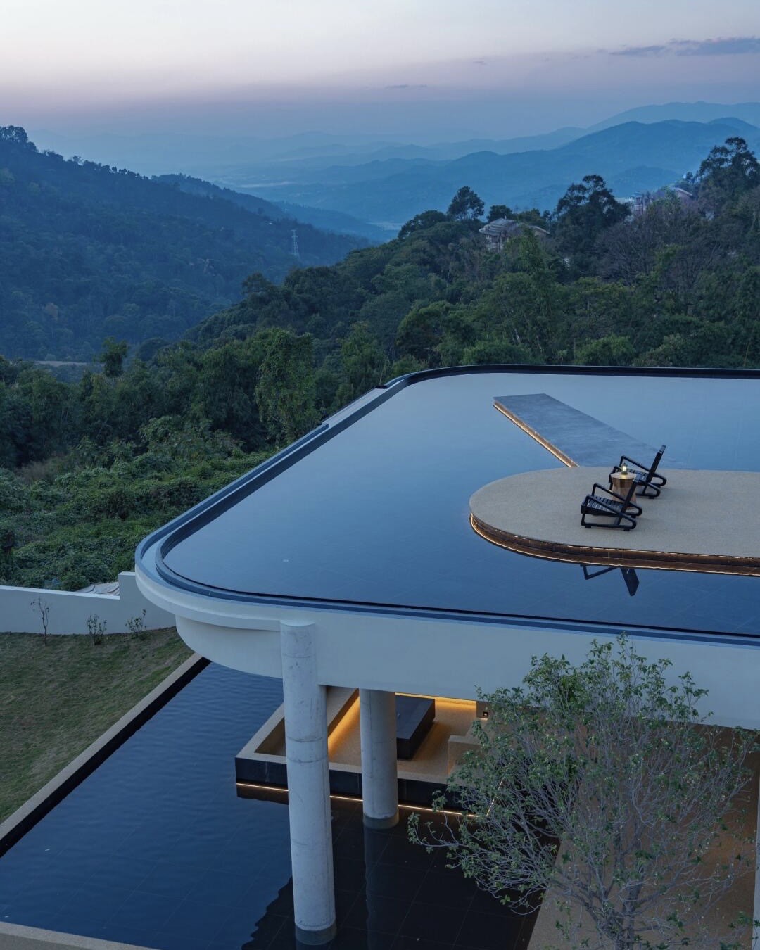 Curved rooftop terrace with loungers above a reflecting pool, overlooking forested hills and hazy mountains at dusk.