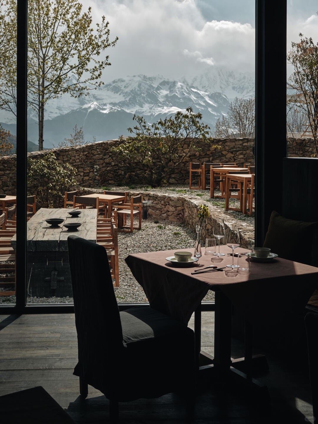 Set dining table by a full-height window looking past a stone-walled terrace to distant snow-capped peaks.