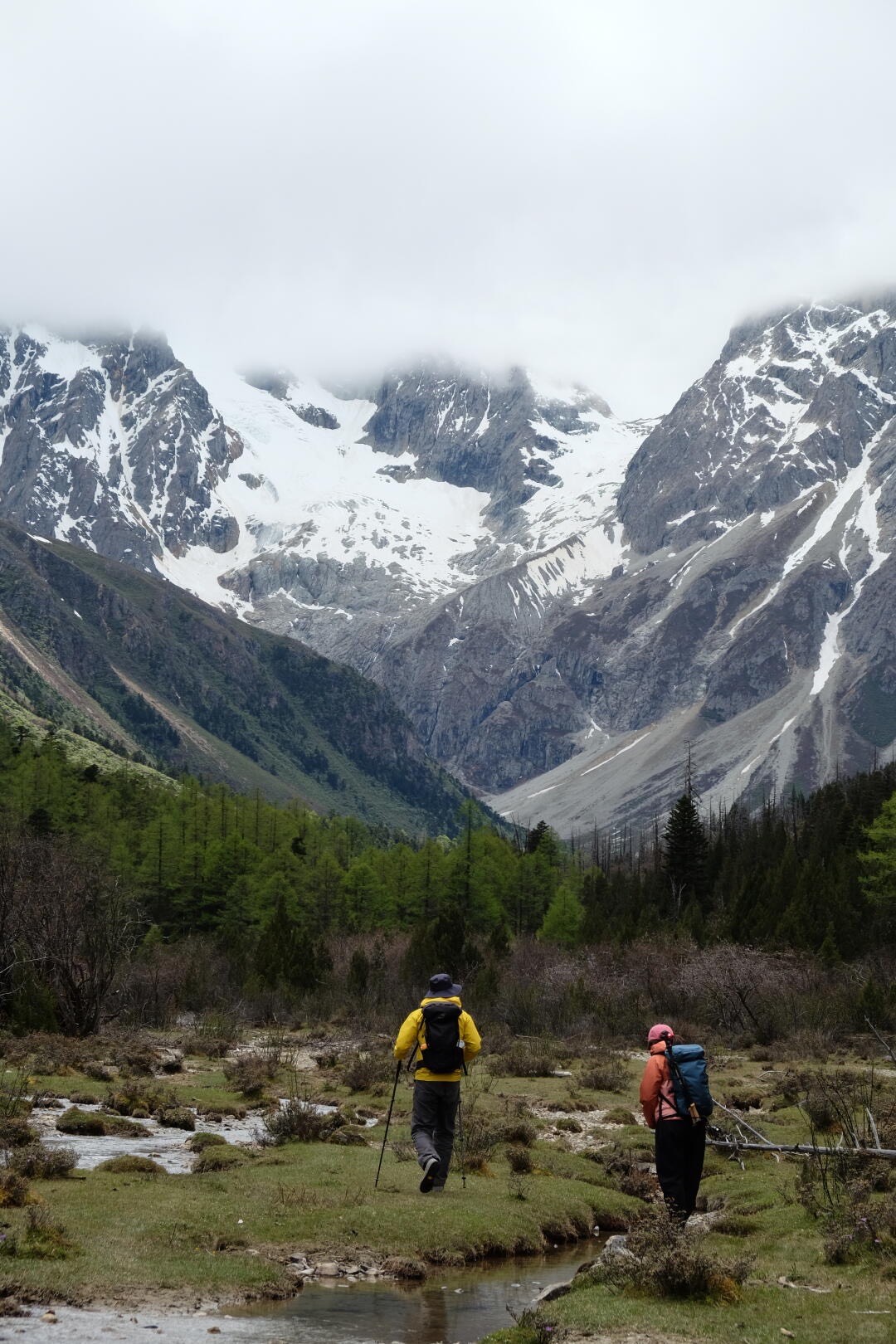 Two backpackers cross a green alpine meadow toward a snow and glacier-clad mountain under low cloud.