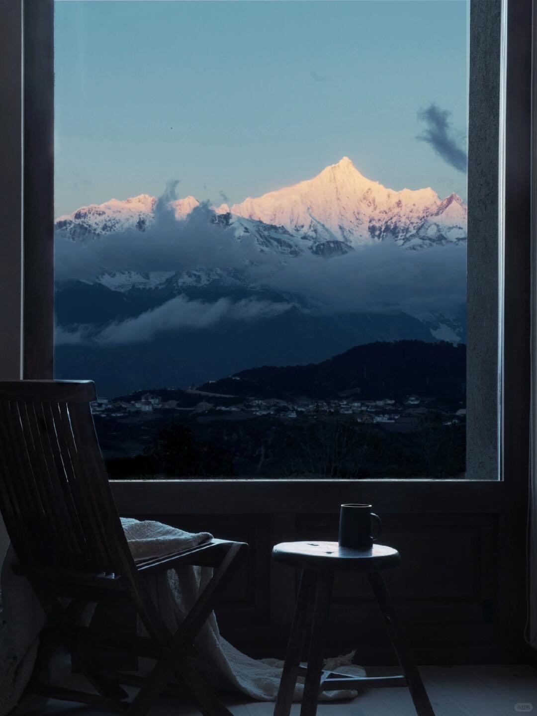 Wooden chair and stool with a mug by a window where sunrise lights a snow-capped peak above a valley town.