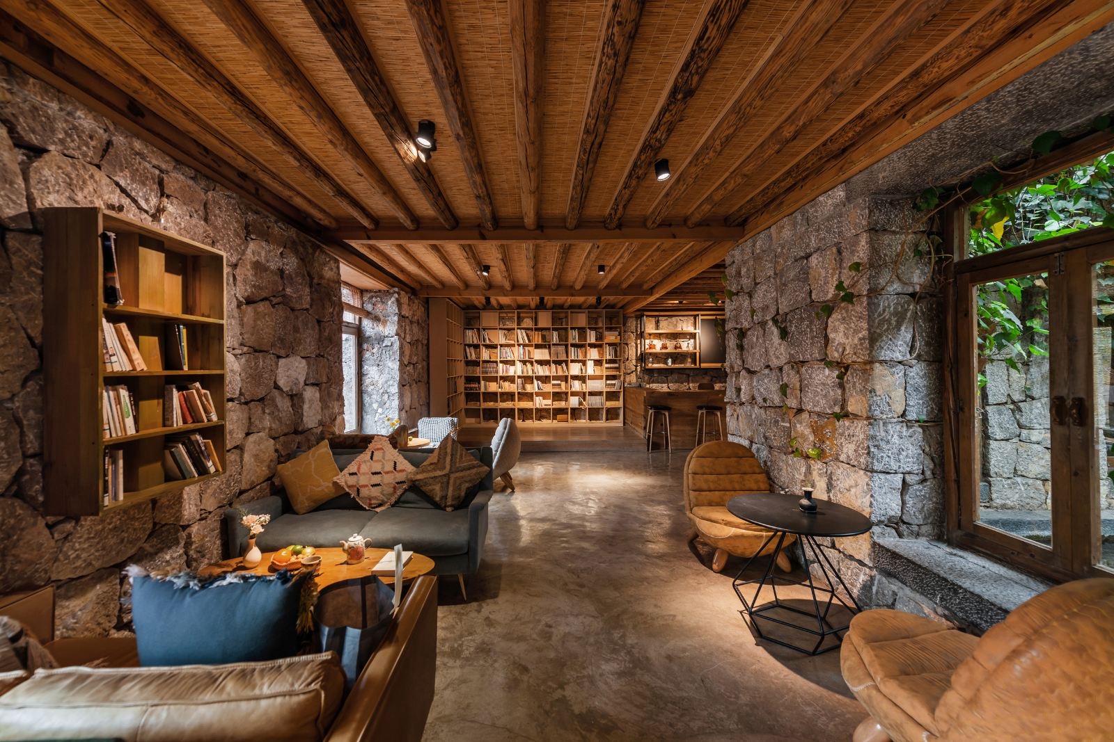 Lanlinge — the library lounge with rough stone walls, exposed timber beams and floor-to-ceiling bookshelves, sofas and armchairs by a garden door