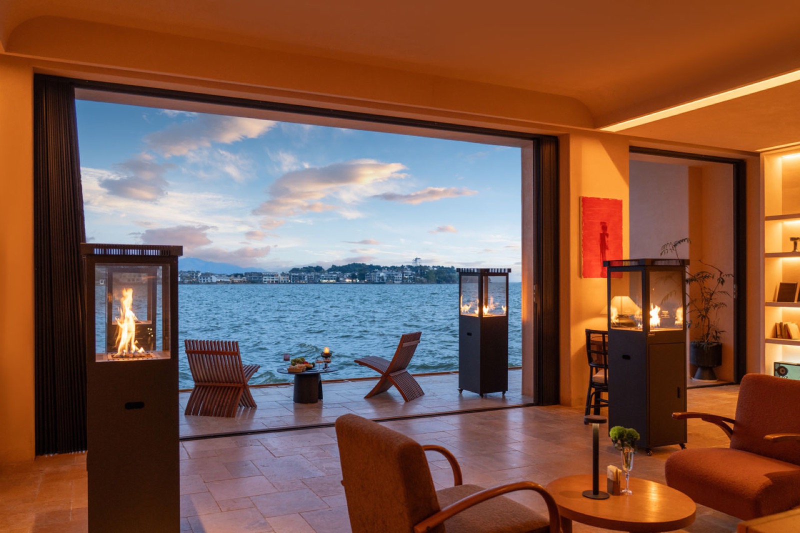 Luna Sea-View Inn — lounge opening through sliding glass onto a travertine terrace on the water, deck chairs and fire pits facing the village across the bay