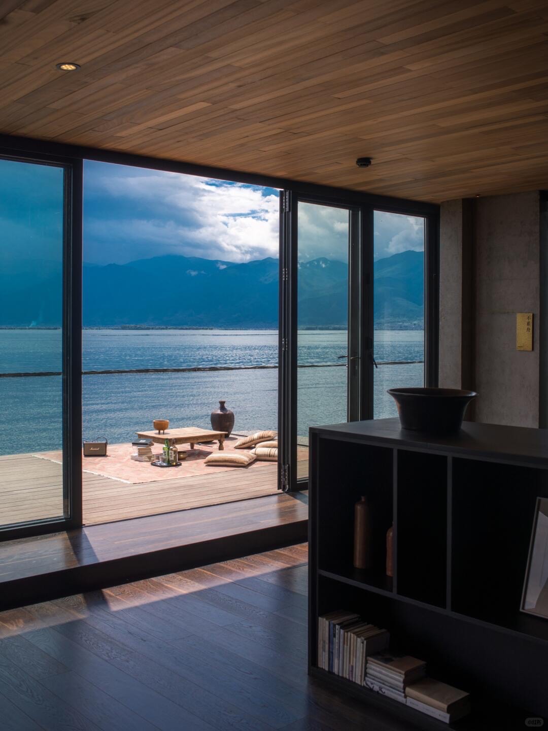 Timber-lined room opening through folding glass doors onto a lakeside deck with low table, cushions and mountains beyond.