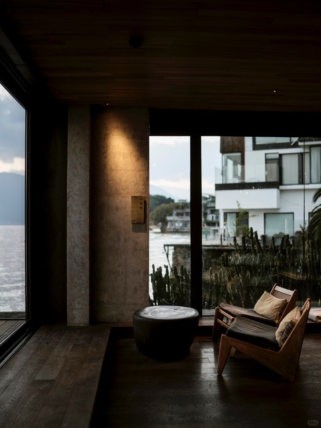 Warm lakeside lounge corner with leather-and-timber chairs and a stone stool facing a lake through glass.